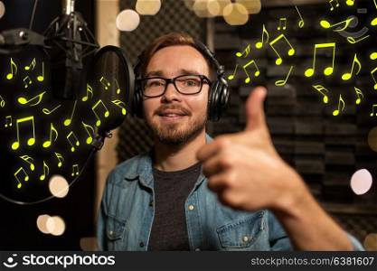 music, show business, people and voice concept - male singer with headphones and microphone singing song at sound recording studio over lights and notes. man with headphones singing at recording studio