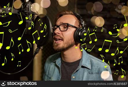 music, show business, people and voice concept - male singer with headphones and microphone singing song at sound recording studio over lights and notes. man with headphones singing at recording studio
