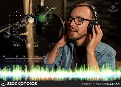 music, show business, people and voice concept - male singer with headphones singing to microphone at sound recording studio. man with headphones singing at recording studio