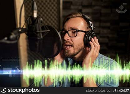 music, show business, people and voice concept - male singer with headphones singing to microphone at sound recording studio. man with headphones singing at recording studio