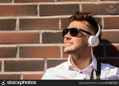 music, technology and people concept - happy smiling young man in headphones and sunglasses over brickwall