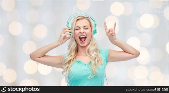music, technology and people concept - happy young woman or teenage girl with headphones singing song and showing rock gesture over holidays lights background