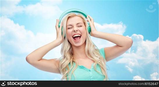 music, technology and people concept - happy young woman or teenage girl with headphones singing song over blue sky and clouds background