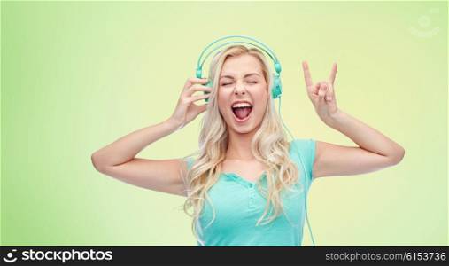music, technology and people concept - happy young woman or teenage girl with headphones singing song and showing rock gesture over green natural background