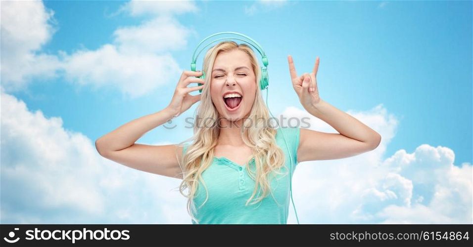 music, technology and people concept - happy young woman or teenage girl with headphones singing song and showing rock gesture over blue sky and clouds background