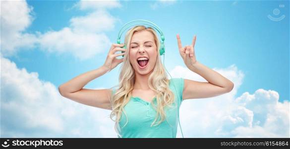 music, technology and people concept - happy young woman or teenage girl with headphones singing song and showing rock gesture over blue sky and clouds background