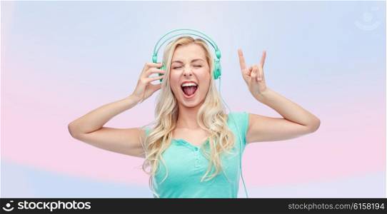 music, technology and people concept - happy young woman or teenage girl with headphones singing song and showing rock gesture over pink background