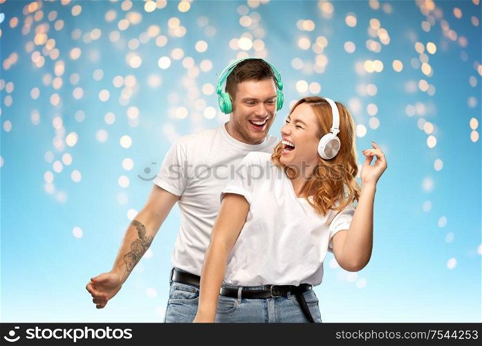 music, technology and people concept - portrait of happy couple in white t-shirts and headphones dancing over holidays lights on blue background. happy couple in headphones dancing
