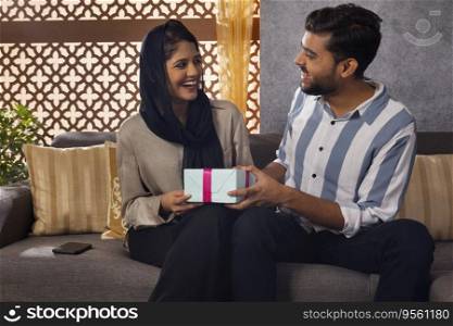 Muslim man giving gift to his happy wife sitting on sofa at home