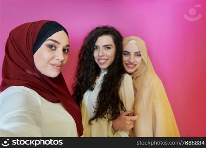 Muslim women taking selfie by mobile phone isolated on pink background. Group of pretty Arab girls in modern fashionable dress using technology