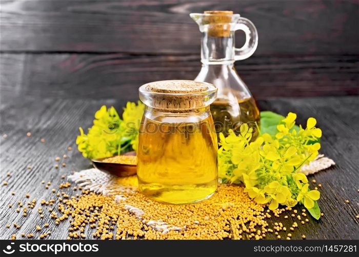 Mustard oil in a glass jar and decanter, mustard grains on a burlap napkin, flowers and leaves on wooden board background