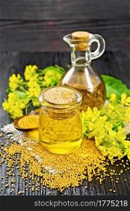Mustard oil in a glass jar and decanter, mustard seeds on burlap, flowers and leaves on dark wooden board