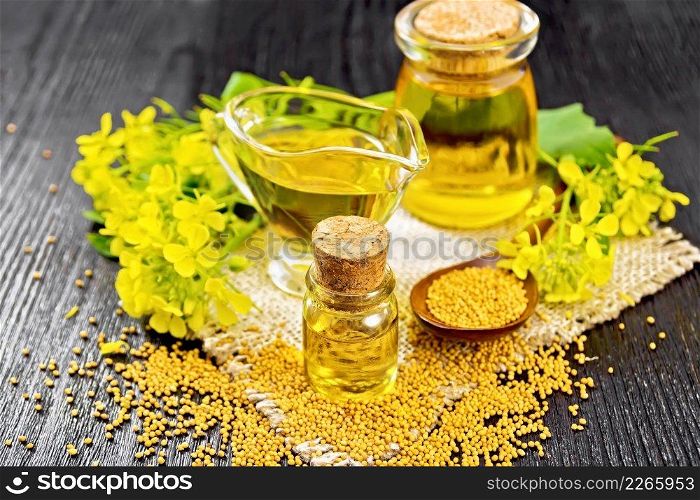 Mustard oil in two glass jars and a sauceboat, grains, leaves and mustard flowers on a burlap napkin on wooden board background