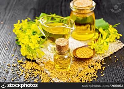 Mustard oil in two glass jars and a sauceboat, grains, leaves and mustard flowers on a burlap napkin on wooden board background