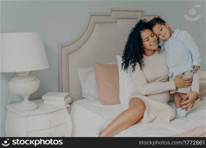 My little boy. Happy pregnant afro american woman in casual white dress with curly hair sitting on bed with her sweet little son and showing strong feelings of love and care. Maternity and pregnancy. Happy pregnant mixed race woman cuddling with her cute little son while relaxing in bedroom
