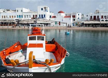 Mykonos town panorama with Greek Orthodox church. Traditional white buildings on embankment and boats on quay. Harbor of Aegean sea, Mykonos island. People walking on bay, Greece