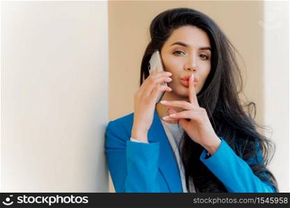 Mysterious brunette woman with makeup, talks with business party, tells secret, makes silent gesture, looks straightly at camera, wears formal apparel. People, communication and conspiracy concept