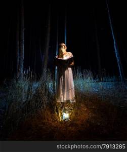 Mysterious shot of beautiful woman in nightgown reading big magical book at dark forest