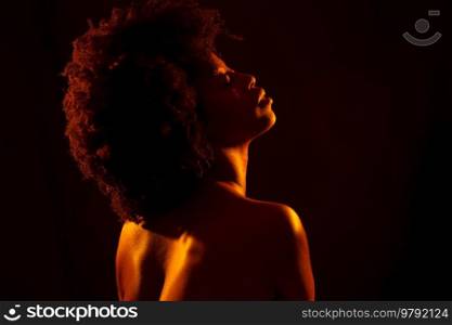 Naked black woman model with Afro hairstyle closing eyes and enjoying bright neon orange light against black background. Nude African American female under orange light
