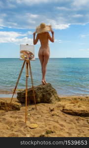naked girl posing in front of an easel with a palette on the seashore
