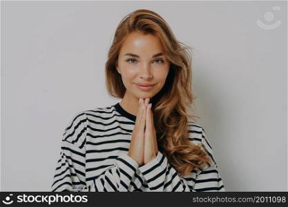 Namaste. Pleased attractive young woman keeps palms pressed together says thank you being very grateful looks polite dressed in casual striped jumper isolated over grey background appreciates offer. Namaste. Pleased attractive young woman keeps palms pressed together says thank you