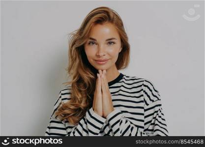 Namaste. Pleased attractive young woman keeps palms pressed together says thank you being very grateful looks polite dressed in casual striped jumper isolated over grey background appreciates offer. Namaste. Pleased attractive young woman keeps palms pressed together says thank you