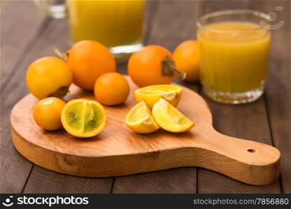 Naranjilla or Lulo fruits (lat. Solanum quitoense) on wooden board with freshly prepared naranjilla juice in the back (Selective Focus, Focus on the naranjilla pieces in the front)