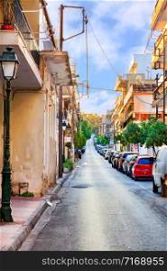 Narrow carriageway against a background of parked cars, a deserted street and bright morning sunlight in blurry spots.. Deserted old narrow street of Loutraki in Greece in the early morning in the rays of the rising summer sun.