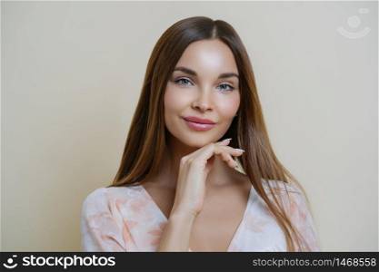 Natural beauty and people concept. Portrait of beautiful dark haired young European woman touches jawline gently, dressed in stylish dress, isolated over beige background, dreams about perfect day