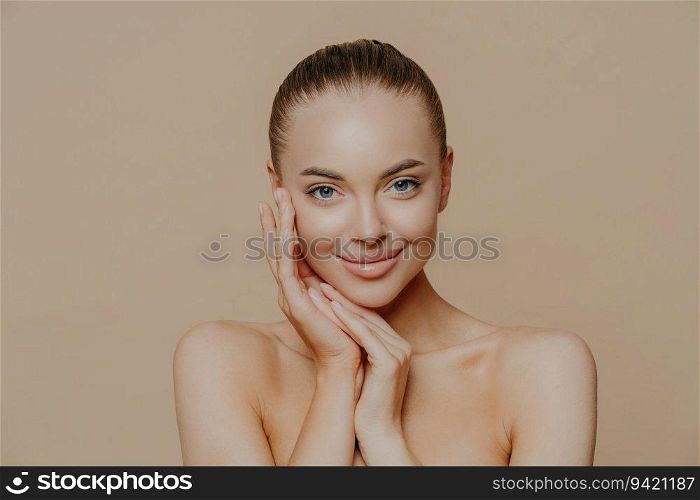 Natural beauty portrait. Fresh skin, gentle smile. Naked pose against beige background. Face touch, minimal makeup.