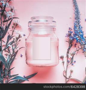 Natural cosmetics jar with pastel pink cream or peeling , herbal leaves and wild flowers, blank label for branding mock-up on pastel background, top view