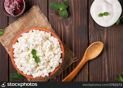 Natural cottage cheese in a traditional clay bowl, next to a wooden spoon, cranberry jam and sour cream in bowls, dark wooden background, top view. Soft curd natural healthy food, wholesome diet food