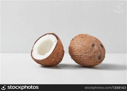 Natural exotic ripe coconut fruit with whole one and half of nut on a duotone light grey background with soft shadows, copy space. Vegan concept.. Half and whole natural organic coconut fruits on a duotone grey background.