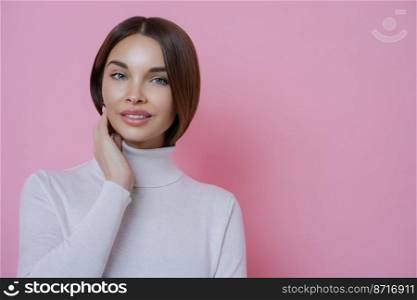 Natural looking young European female model smiles gently at camera, has well cared dark hair, dressed in casual white poloneck, poses against pink background, blank space for your information