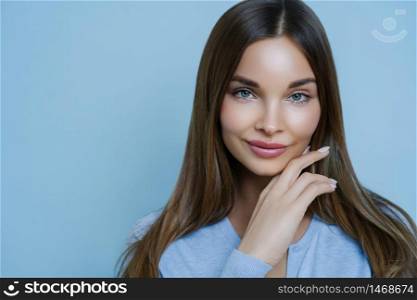 Natural magnetic young woman with fresh clean skin, has attractive facial features, gazes sensually at camera, feels beautiful and appealing, touches jawline, dressed in casual jumper isolated on blue