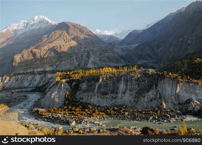 Natural sunlight shining at colorful trees and river against snow capped mountain peaks in Karakoram range. Hunza Nagar Valley in autumn season. Gilgit Baltistan, Pakistan.