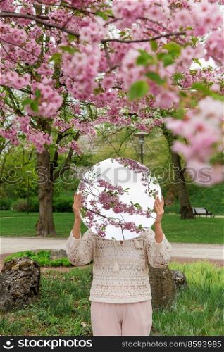 nature and people concept - woman holding round mirror with cherry tree reflection in spring garden or park. woman with cherry tree reflection in round mirror