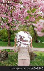 nature and people concept - woman holding round mirror with cherry tree reflection in spring garden or park. woman with cherry tree reflection in round mirror