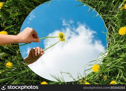 nature concept - hand with dandelion touching sky reflection in round mirror on summer field. hand with dandelion and sky reflection in mirror