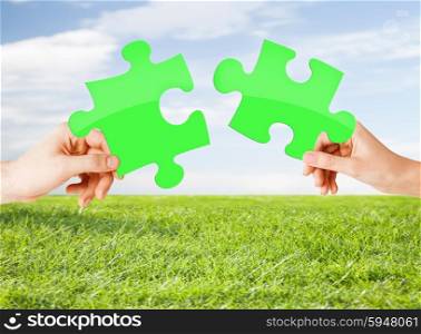 nature, ecology, energy saving, people and environment concept - close up of couple hands trying to connect green puzzle pieces over blue sky and grass background
