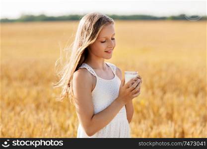 nature, healthy eating and organic concept - smiling young girl holding glass of milk on cereal field in summer. girl with glass of milk on cereal field