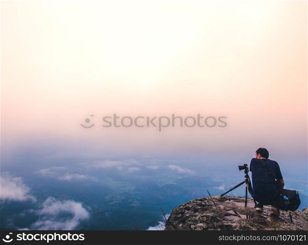 Nature photographer takes photos with camera on tripod at cliff. Dreamy foggy landscape. Morning sunrise blue misty in beautiful valley below.