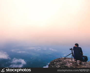 Nature photographer takes photos with camera on tripod at cliff. Dreamy foggy landscape. Morning sunrise blue misty in beautiful valley below.