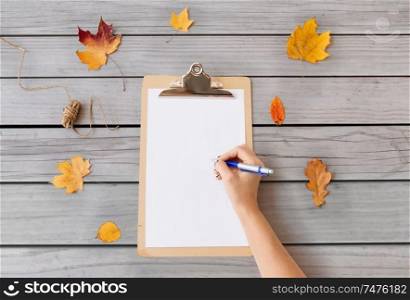 nature, season and botany concept - hand with pen and blank white paper on clipboard and autumn leaves on gray wooden boards background. hand writing on white paper on clipboard in autumn