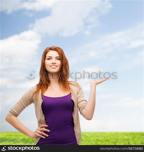 nature, summer, advertising and people concept - smiling teenage girl in casual clothes holding something on her palm over blue sky and grass background