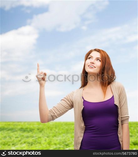 nature, summer, gesture and people concept - smiling teenage girl in casual clothes pointing finger up over blue sky and grass background