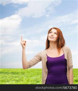 nature, summer, gesture and people concept - smiling teenage girl in casual clothes pointing finger up over blue sky and grass background