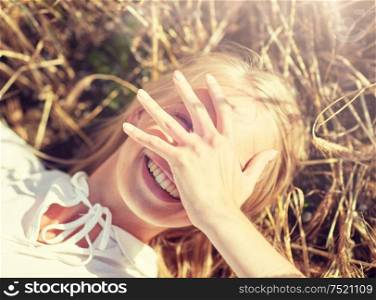 nature, summer holidays, vacation and people concept - close up of happy young woman lying on cereal field and covering face by hand. happy young woman lying on cereal field