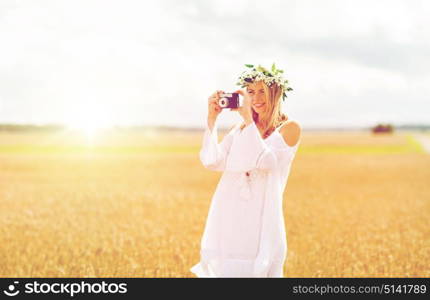 nature, summer holidays, vacation and people concept - happy woman in wreath of flowers taking picture with film camera outdoors. happy woman with film camera in wreath of flowers