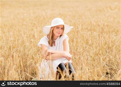 nature, summer holidays, vacation and people concept - happy young woman in white dress, rubber boots and sun hat sitting on cereal field